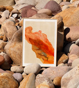 A framed abstract Maison Charlot art poster featuring flowing orange and red brushstrokes on a cream background, displayed upright on a bed of smooth river stones.