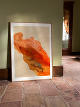 A framed abstract art print leaning against a sage green wall on a terracotta tiled floor. The artwork features flowing, organic shapes in vibrant orange, red, and soft beige tones.